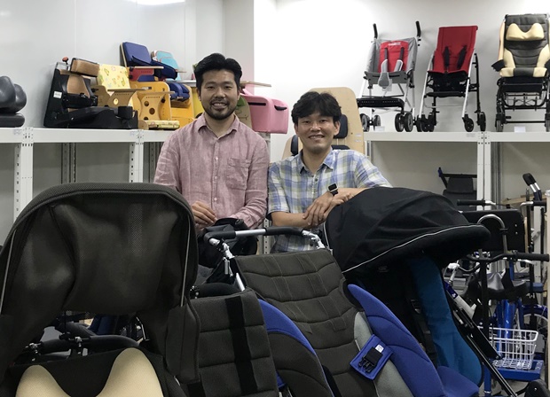 장애아동용 유모차 뒤에서 포즈를 취하고 있는 문상진(좌), 강용원(우) 센터장. 아동이 성장함에 따라 변하는 신체 크기에 맞는 보조기기가 다양하게 전시돼 있다.